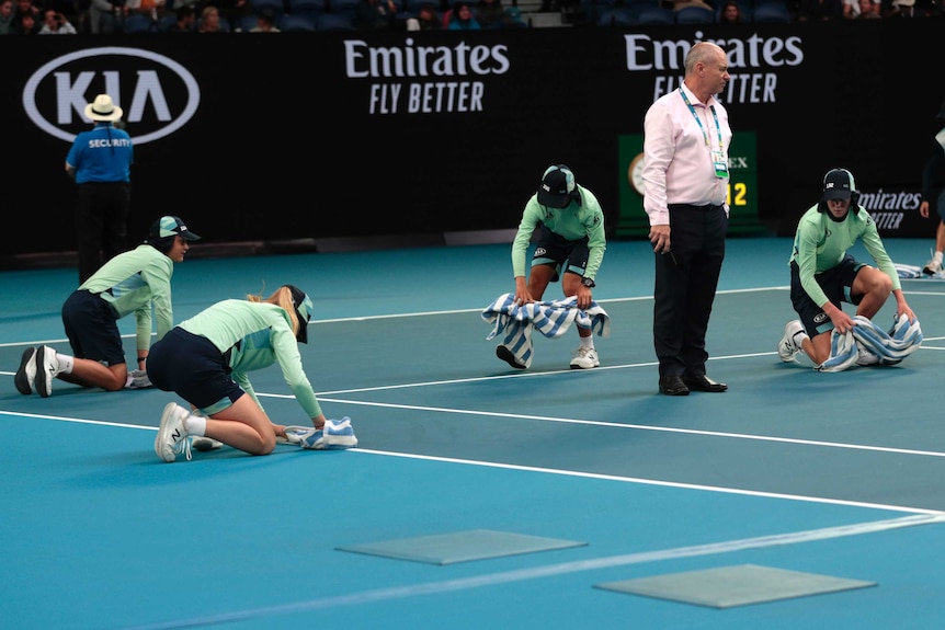 Ball kids can be seen on their knees drying a court with towels whilst a man in a shirt stands in the middle of them