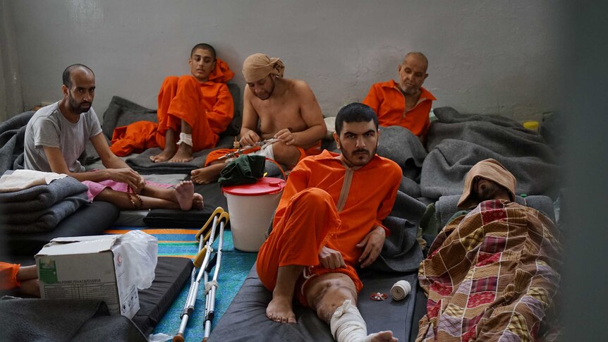 Men dressed in orange prison suits sit in a crowded cell.