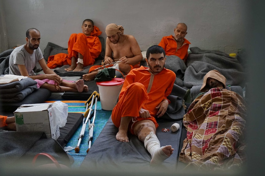 Men dressed in orange prison suits sit in a crowded cell.