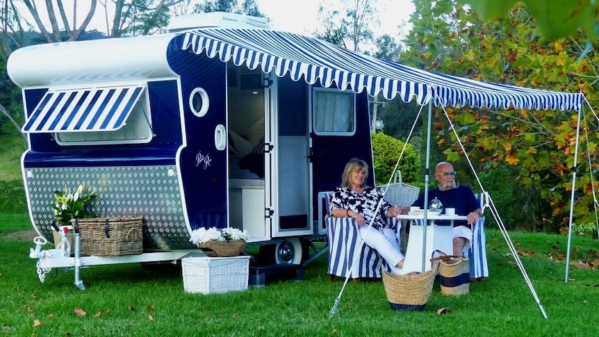 A couple sits outside under the striped awning of their vintage caravan.