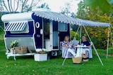 A couple sits outside under the striped awning of their vintage caravan.