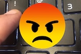 An angry-face emoji superimposed over a close up image of a finger typing an exclamation mark on a keyboard.