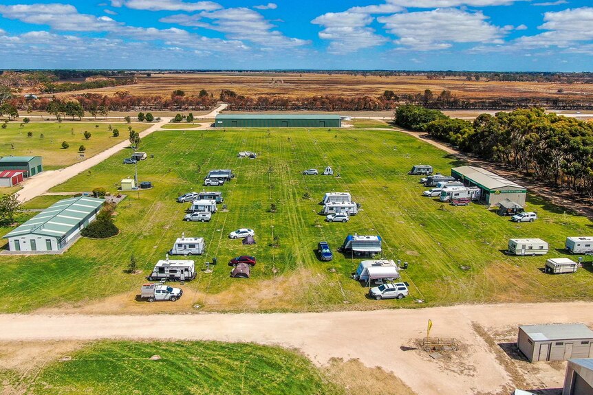 An aerial photo of 20-odd caravans stretched out over a large grass field under a bright blue sky.