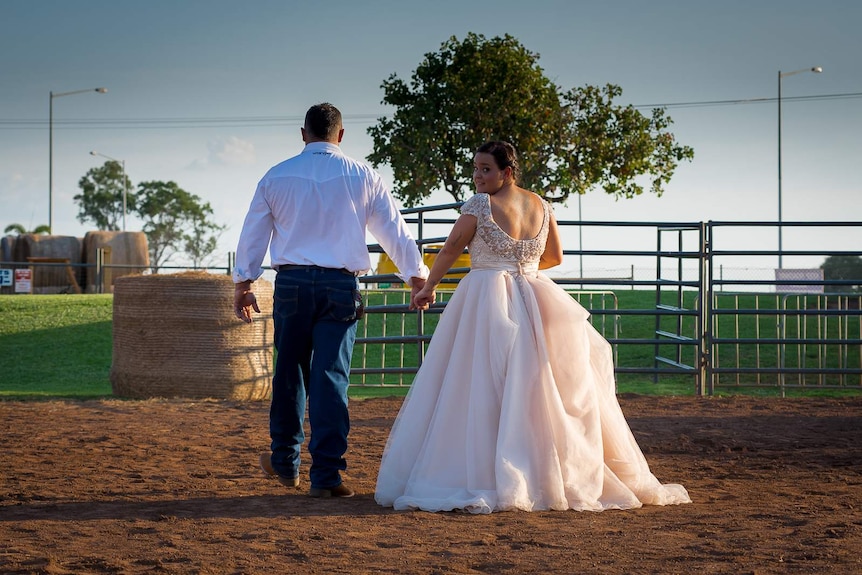 A newly married couple walk through the dirt and dust of a rodeo ring towards the exit, as the bride looks over her shoulder.