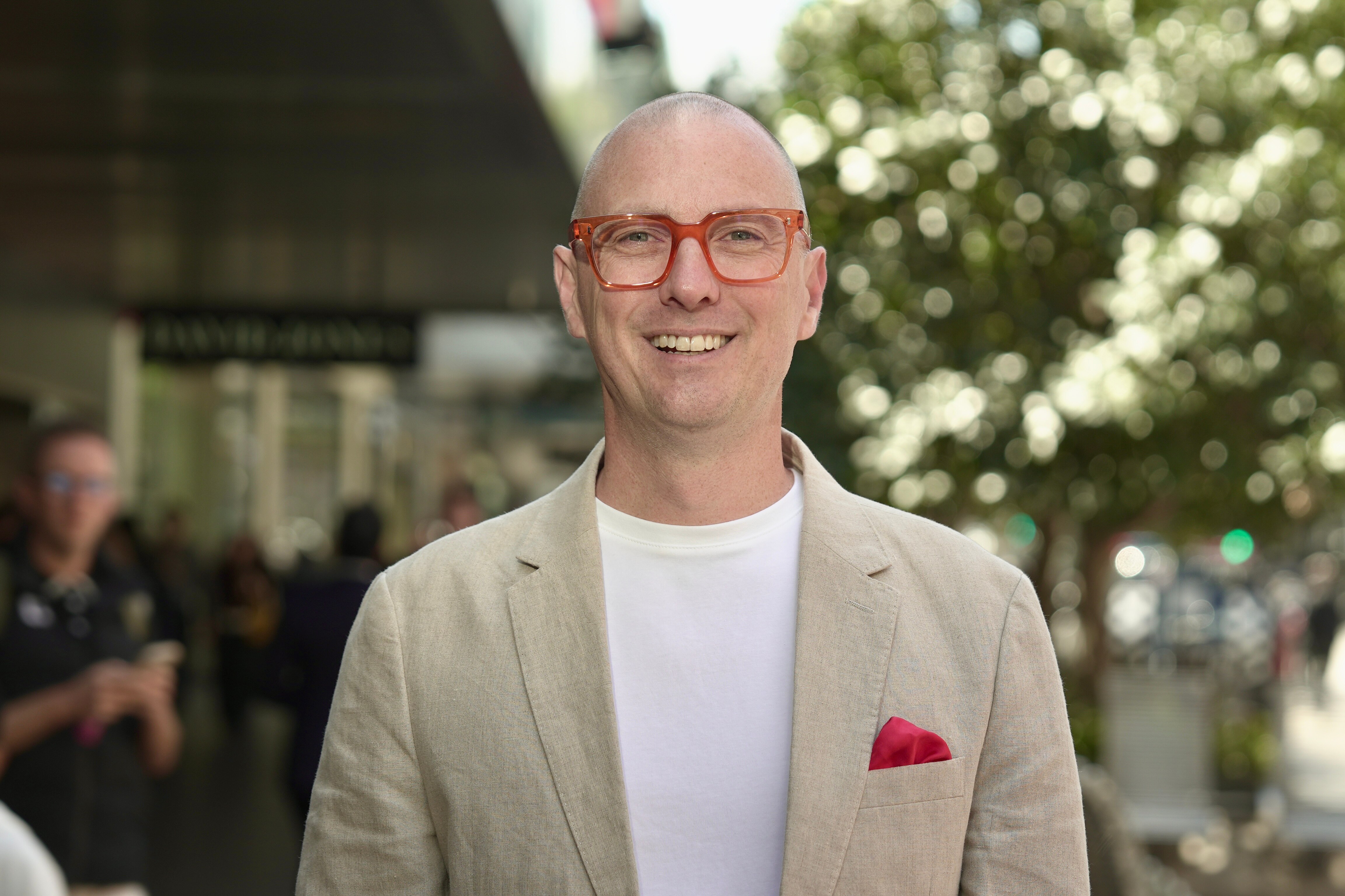 O sorridente James é careca, usa óculos de plástico com armação vermelha, uma jaqueta de linho creme, uma camiseta branca, um lenço de bolso vermelho e um fundo desfocado.