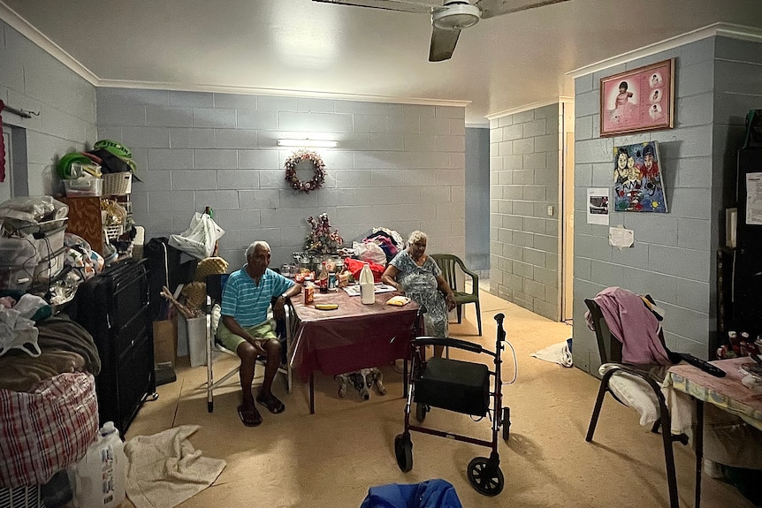 Denise's elderly parents sit at a dining table, lining one wall of the room is storage and containers of clothes.