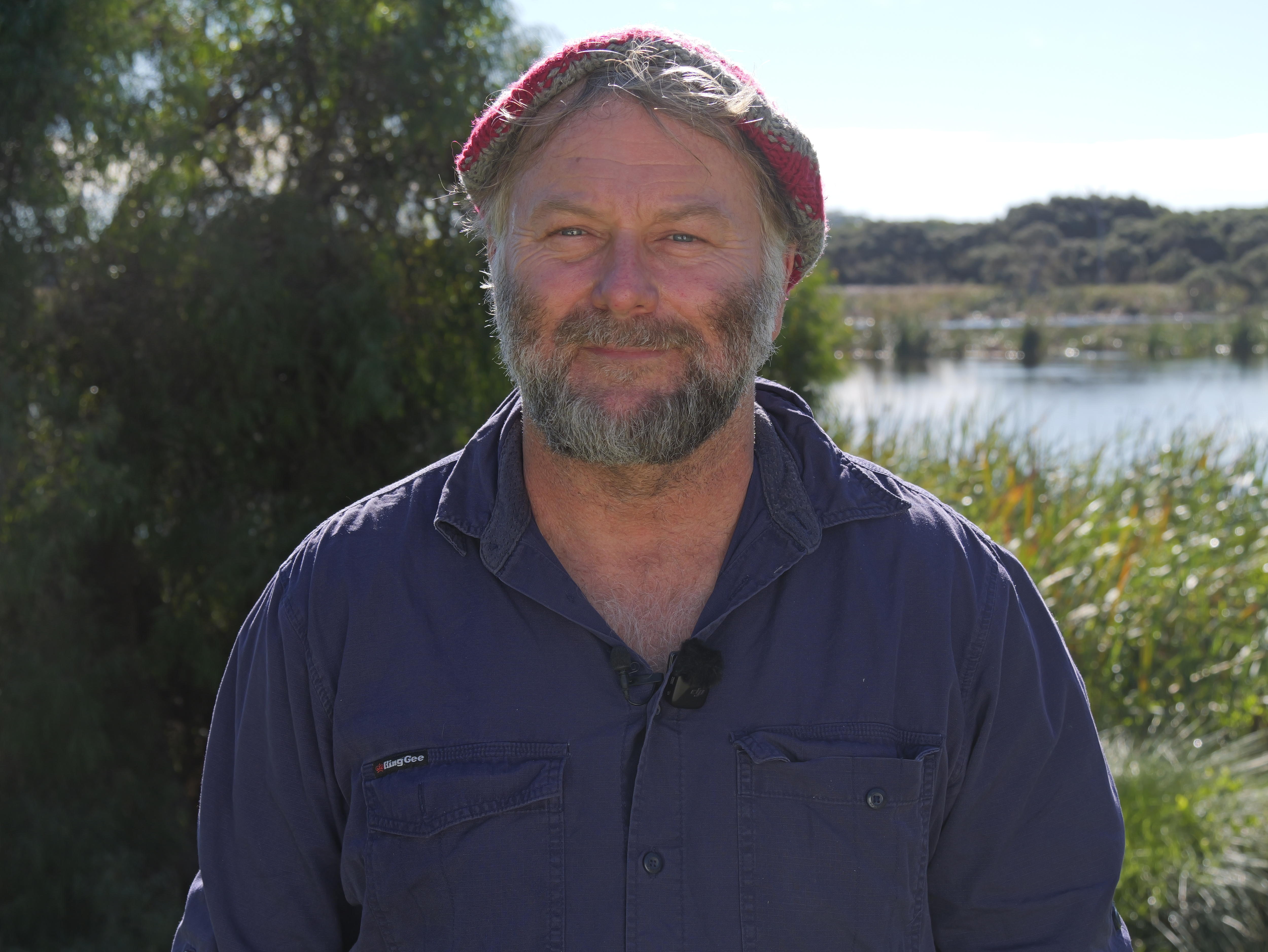 Tim Gamblin wears a blue shirt and a red and grey knitted hat. The wetlands in the background.