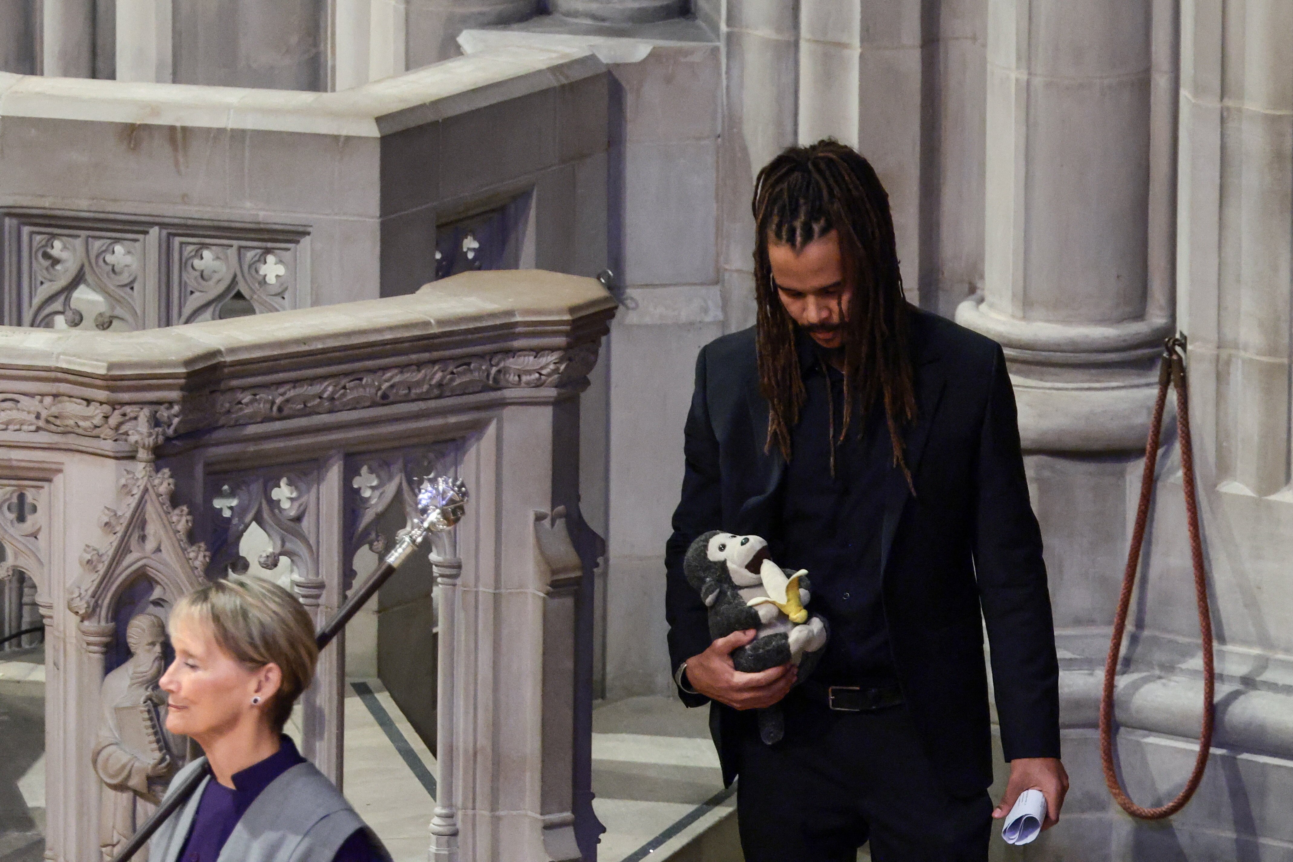 A young man with dreadlocks wears a dark outfit as he walks down a set of stone stairs in a cathedral.