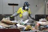 A man wearing a cap, mask and goggles stands at a conveyor belt sifting rubbish.