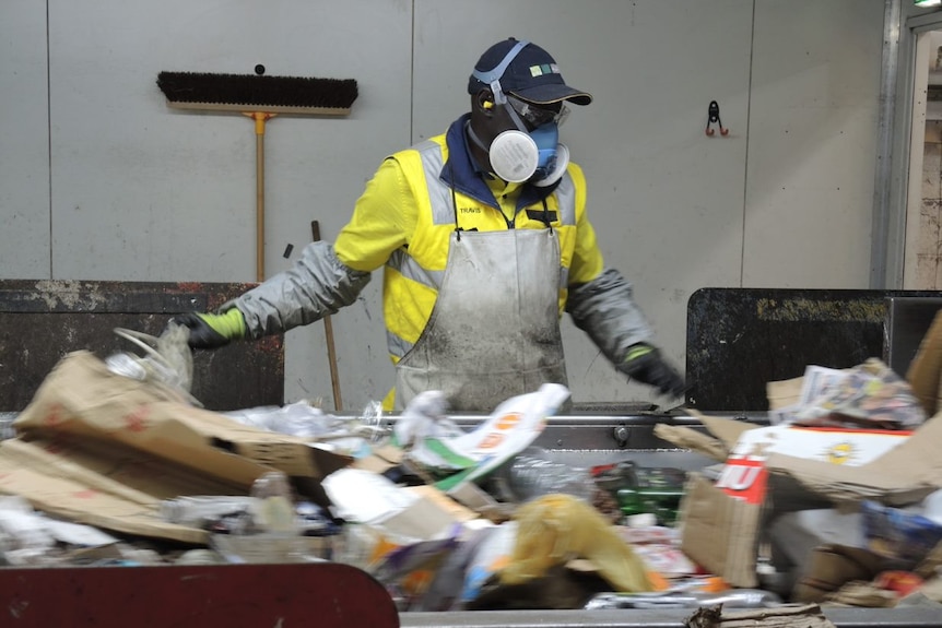 A man wearing a cap, mask and goggles stands at a conveyor belt sifting rubbish.