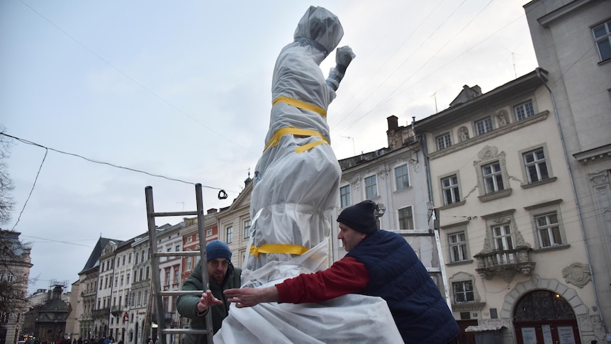 Two men with a ladder wrapping an outdoor statue using tape and fabric.