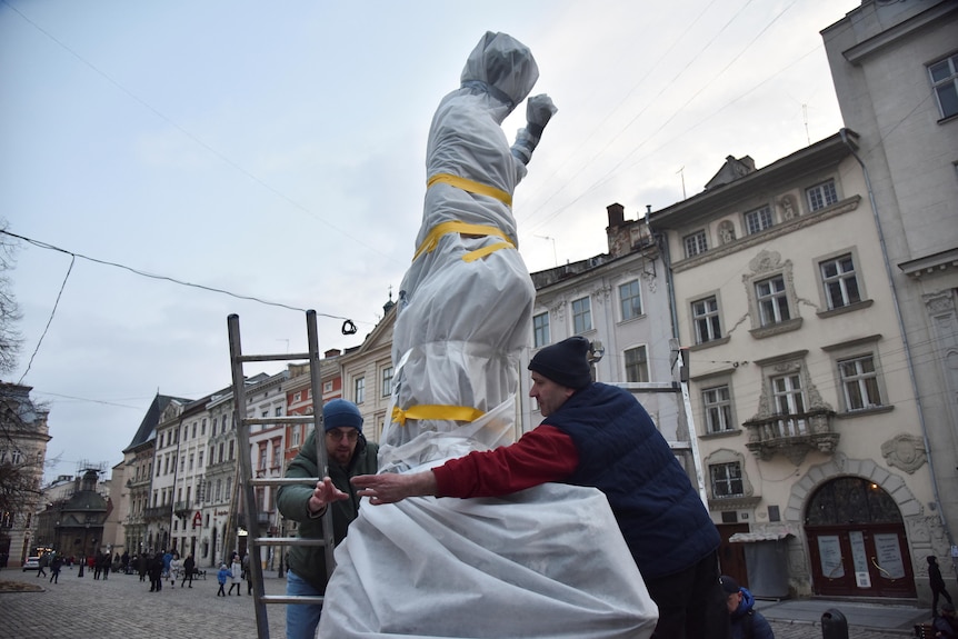 Two men with a ladder wrapping an outdoor statue using tape and fabric.
