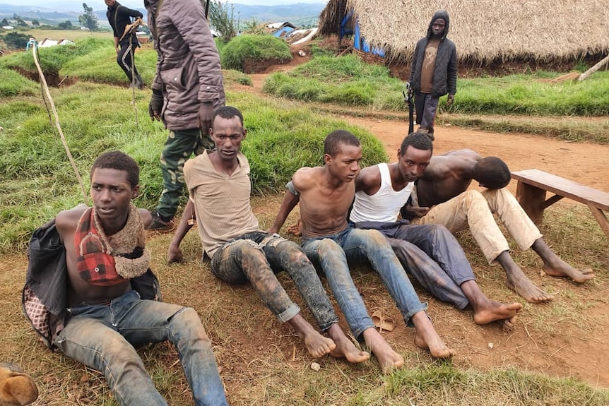 Men sit on the ground while other men, some armed with guns, raid their village. 