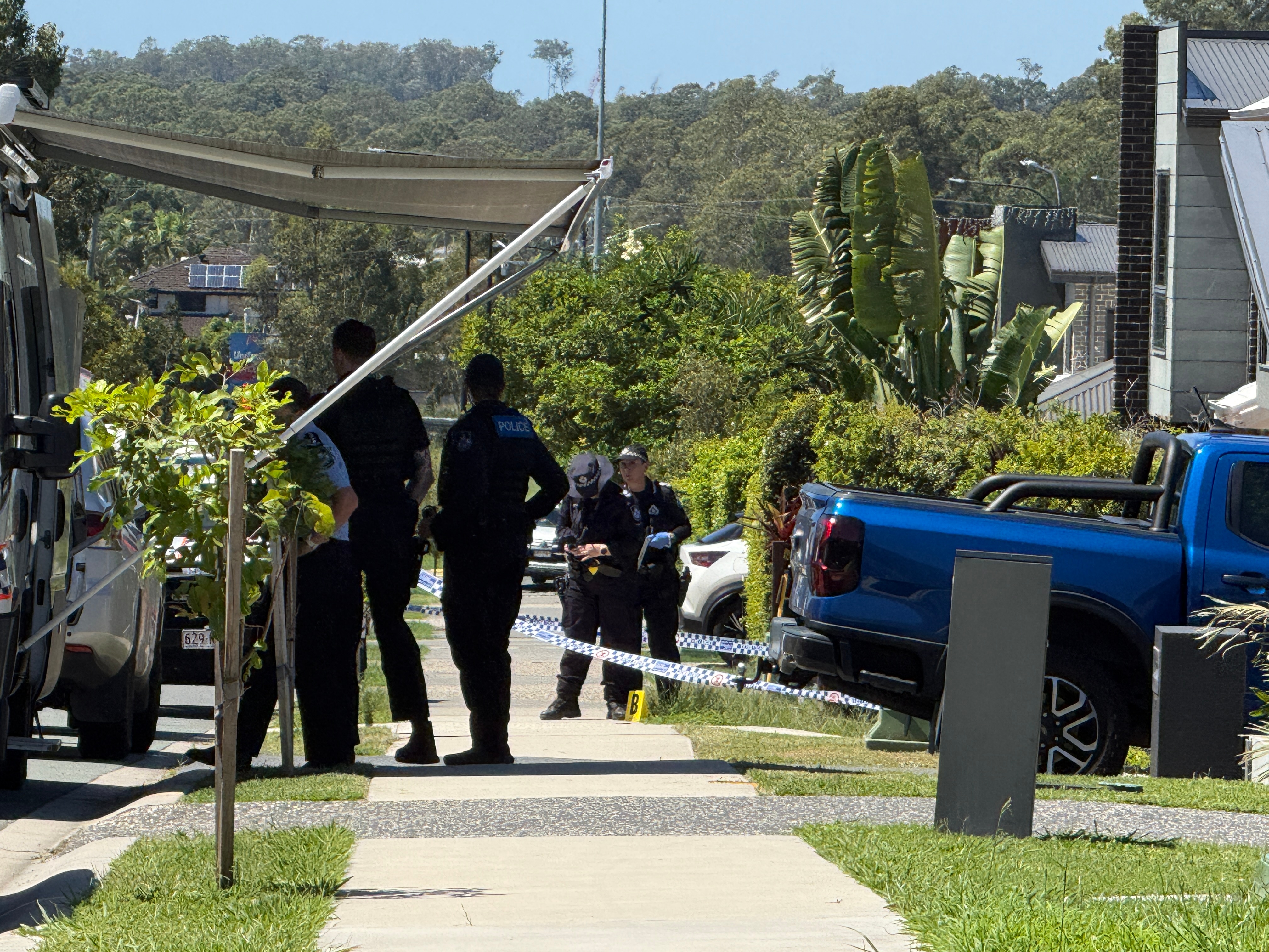 Police officers stand on the footpath outside houses next to a police van with an awning.
