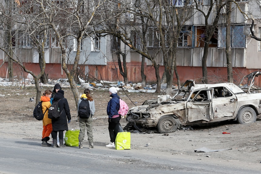 five people with backpacks and bags stand near a burnt-out car in front of a damaged building in the mariupol warzone