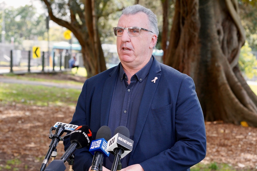 Man in blazer stands in front of microphones with a tree behind him