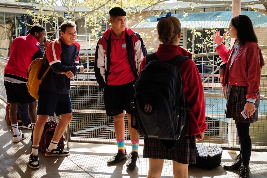 Three teenage boys and two girls in school uniforms stand on a balcony chatting with classrooms in the background