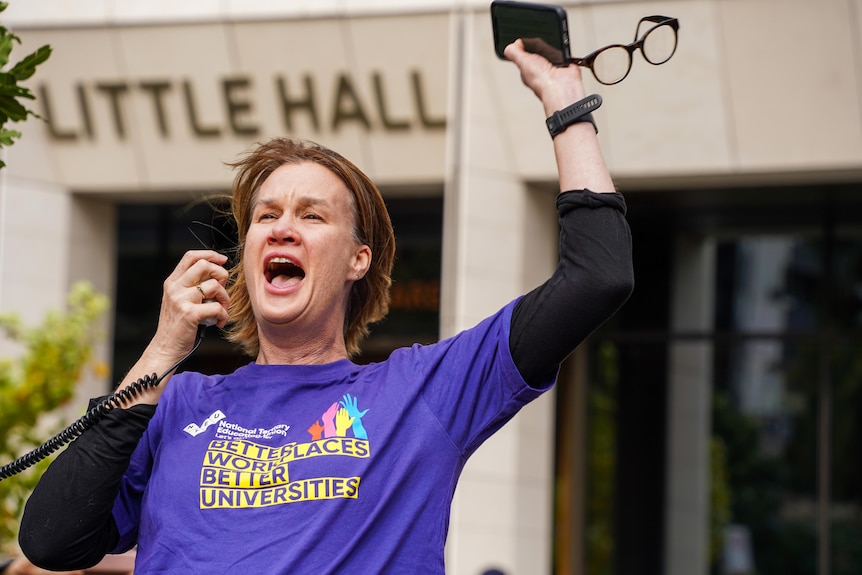 Alison raises an arm in the air as she speaks at a protest, wearing a shirt which says "better workplaces, better universities".