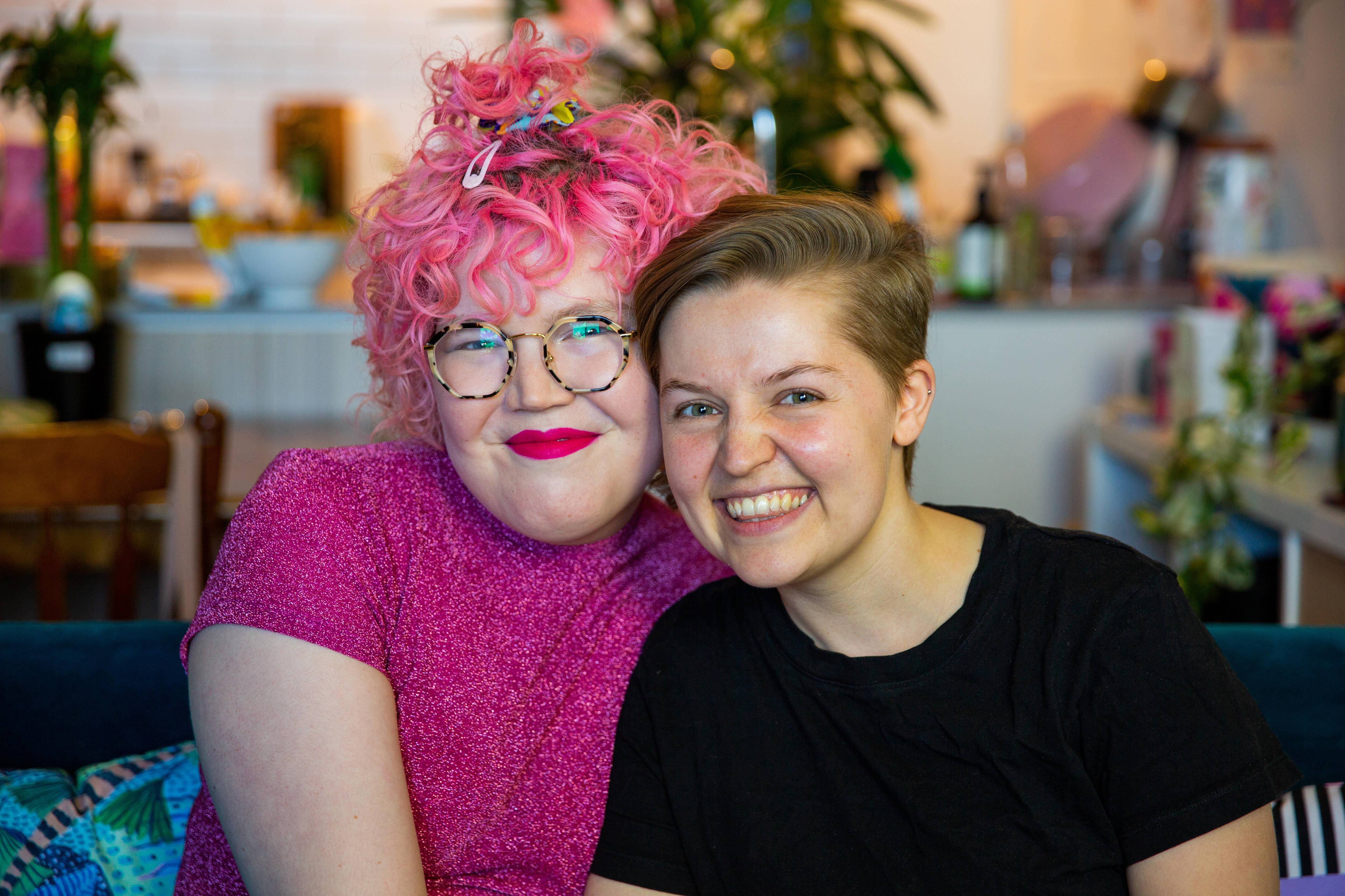 Ivy McGowan (left) sits next to Yoey Maxwell on a sofa in their colourful apartment. They both look at the camera and smile. 
