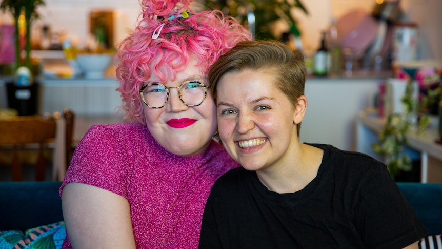 Ivy McGowan (left) sits next to Yoey Maxwell on a sofa in their colourful apartment. They both look at the camera and smile. 