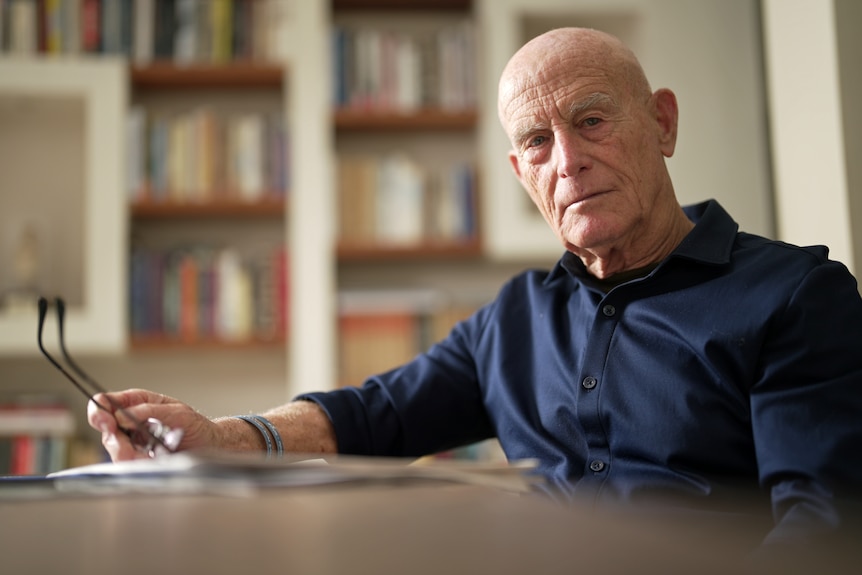 A man sitting at a desk holding glasses looks at the camera. Behind him are bookshelves.