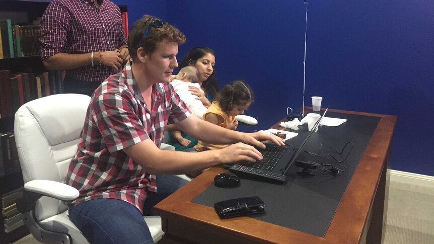 Logan Turnbull types on a computer with his family by his side and a helper watching over.