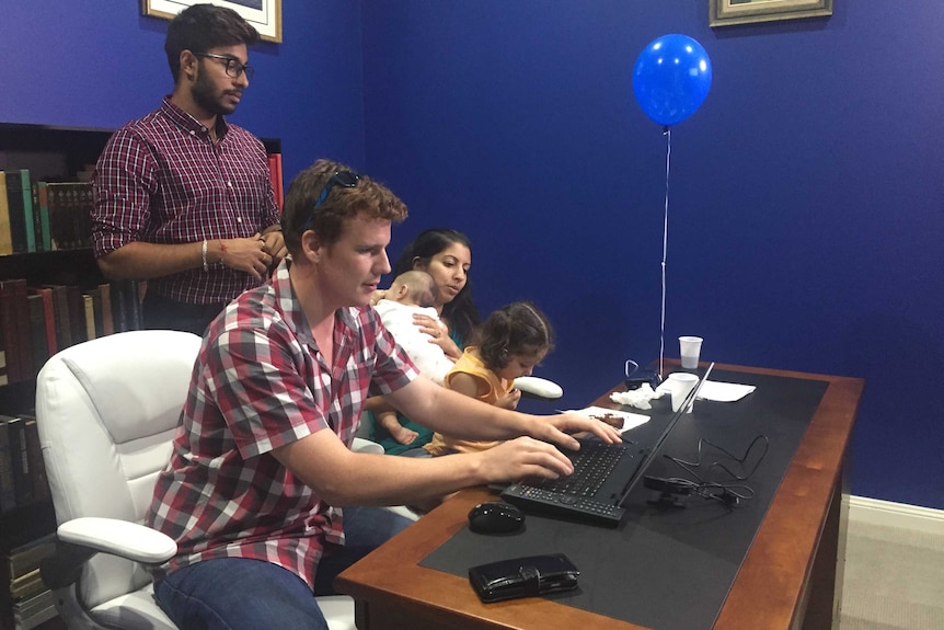 Logan Turnbull types on a computer with his family by his side and a helper watching over.