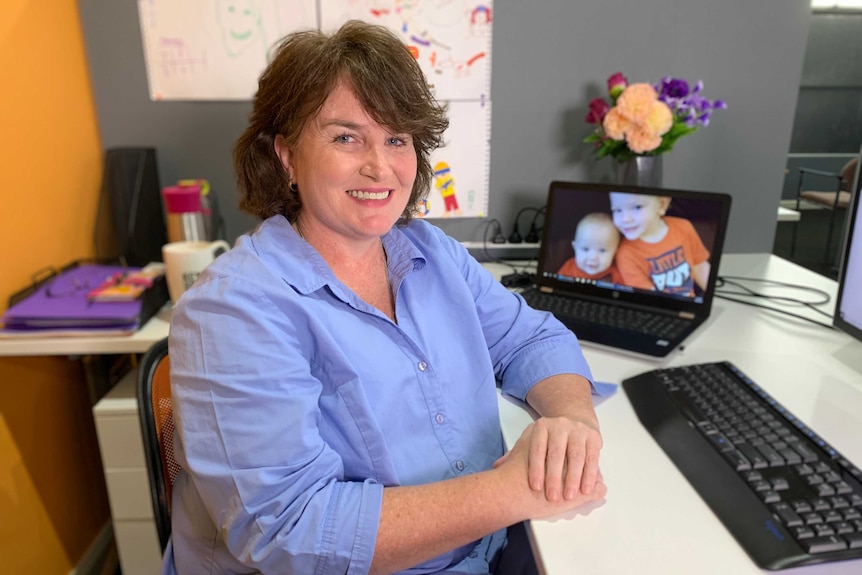 A woman at a desk with a laptop behind with a screensaver of a baby and a toddler