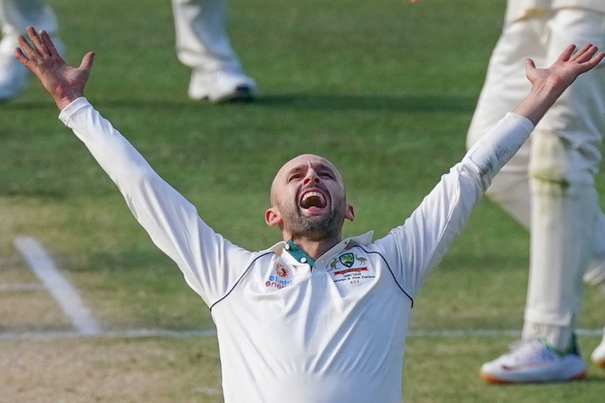 A bald man throws his hands in the air as he shouts while standing on a cricket pitch with other players behind him.