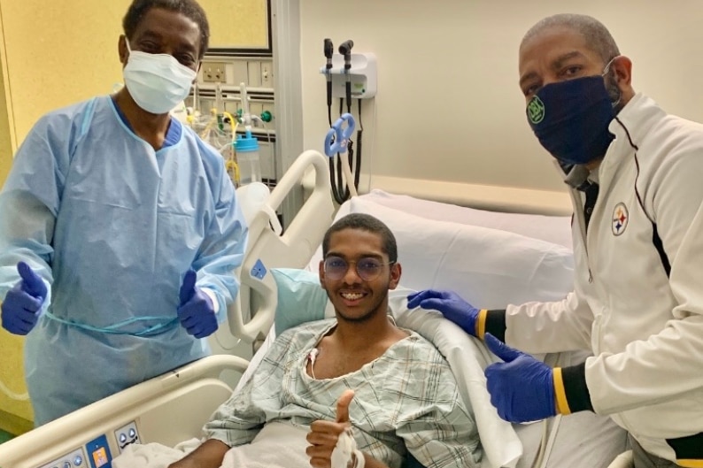 David Vargas lies in a hospital bed with Dr George Ofori-Amanfo (left) and his father (right) standing next to him.