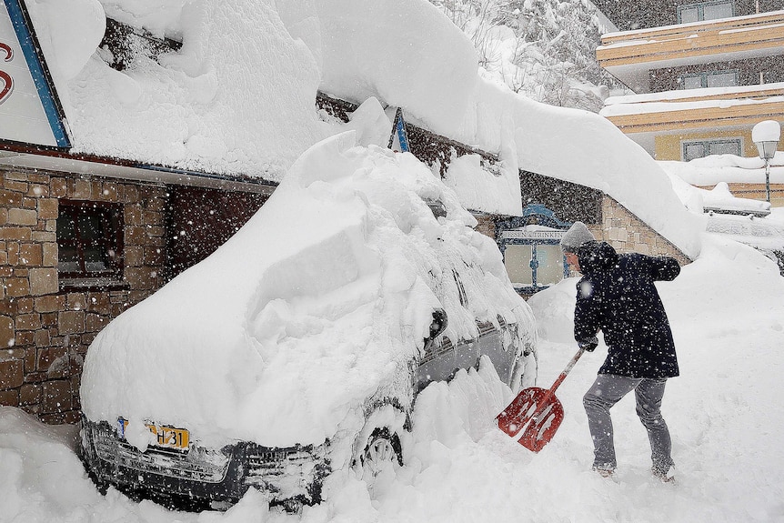 A man shovels snow from his car, parked next to a building and streets heaped with snow as it continues to fall.