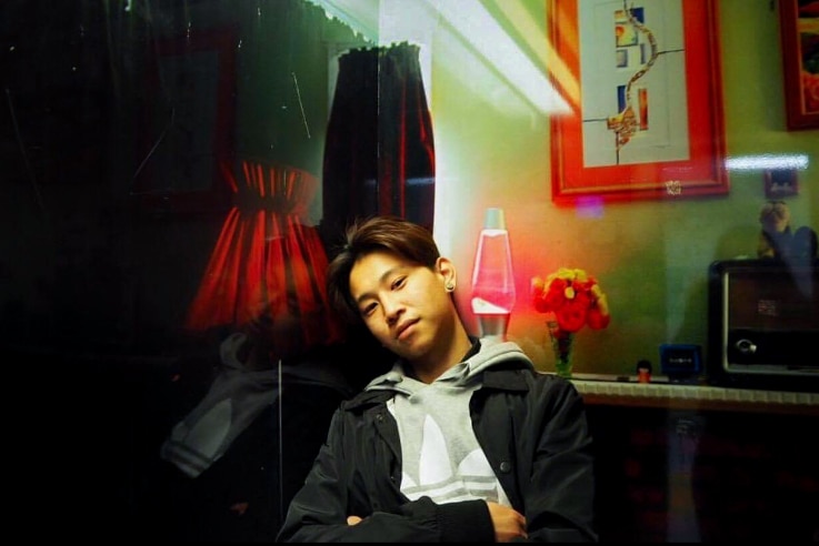 Dillon Wu, wearing grey hoodie and black jacket, sits arms folded in a reflective living room.