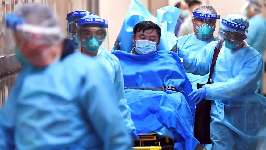 A masked patient is transferred on a stretcher by masked medical staff wearing blue overalls.