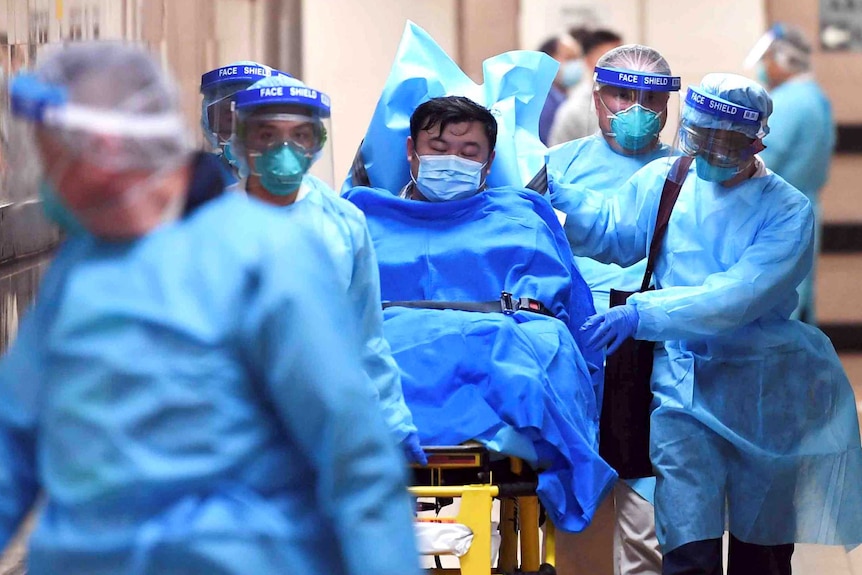 A masked patient is transferred on a stretcher by masked medical staff wearing blue overalls.