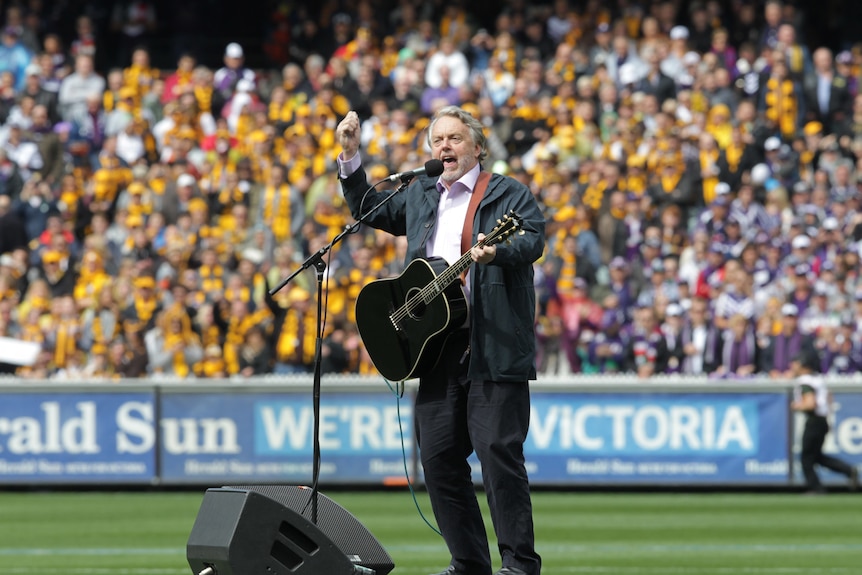 A man with a guitar puts his hand in the air as he stands in the middle of the MCG and sings at a packed AFL grand final.
