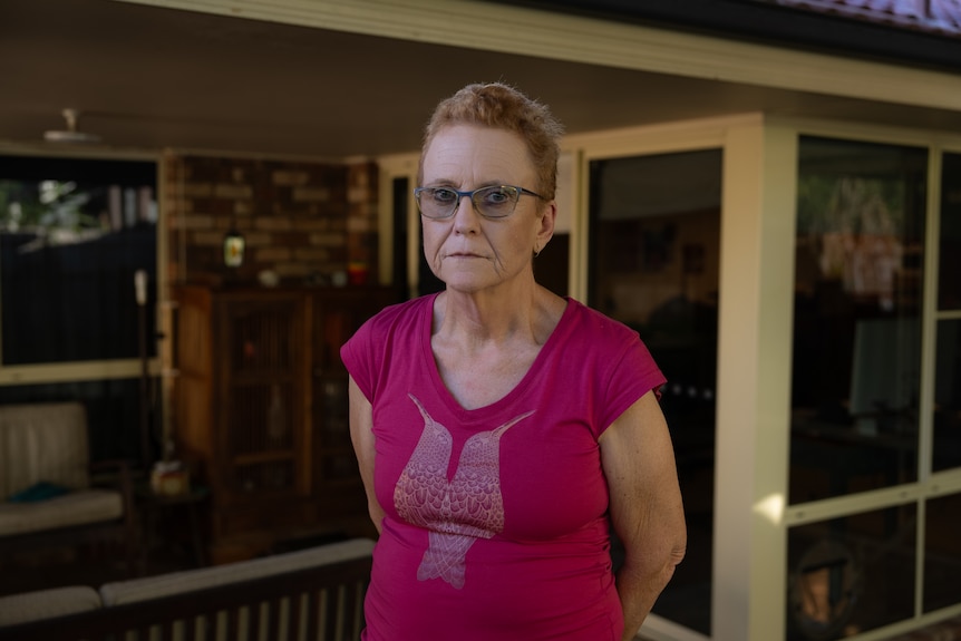 A woman wearing glasses stands outside, looking into camera with a serious expression.