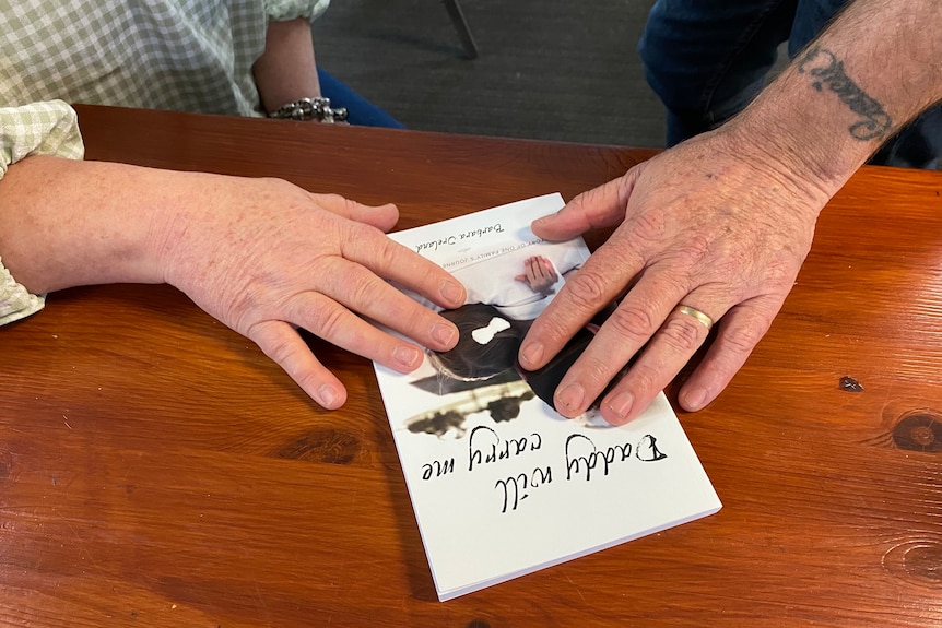 A woman and a man's hands resting on a white book called 'Daddy will carry me' sitting on a table 