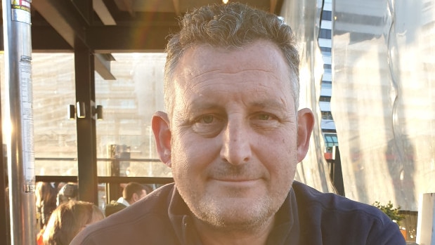 A smiling man sits at an outdoor table with a glass of beer.