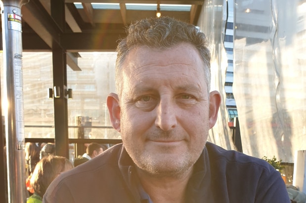 A smiling man sits at an outdoor table with a glass of beer.