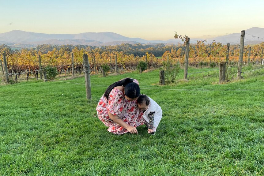 A woman and child huddled and looking at something in the green grass with vinyards and hills in the background.