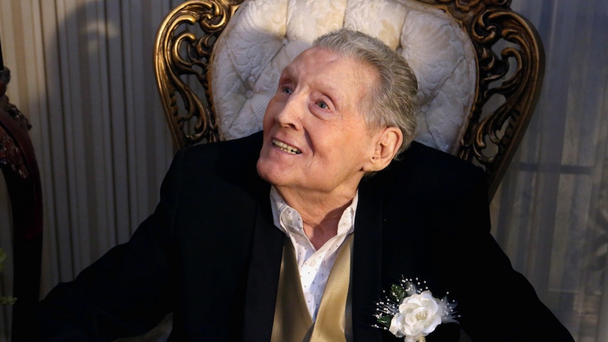 jerry lee lewis sits on a chair smiling wearing a black suit, gold waistcoat and white flower pin