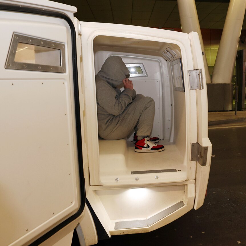 a man wearing a grey track suit with a hoodie covering his face sits inside a police van 