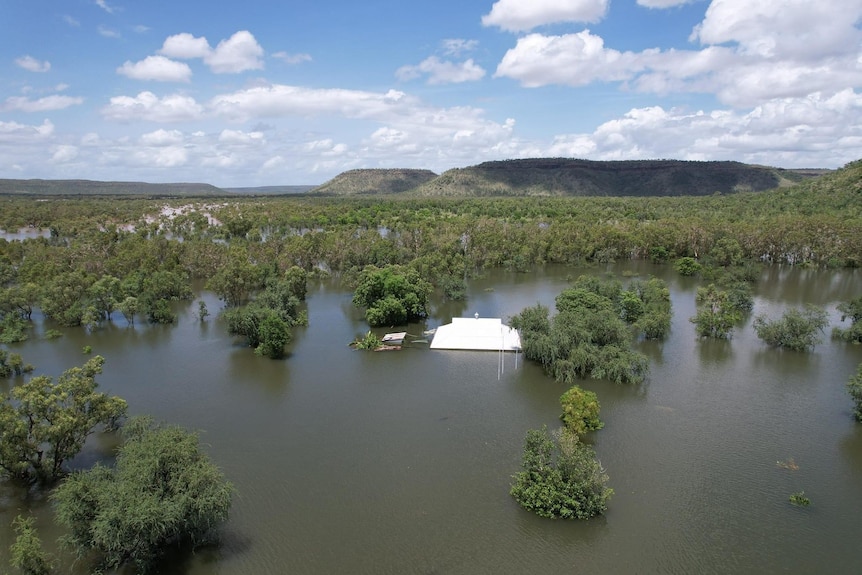 A white roof and the tops of trees peak out of green-brown floodwater, with mountains in the background.