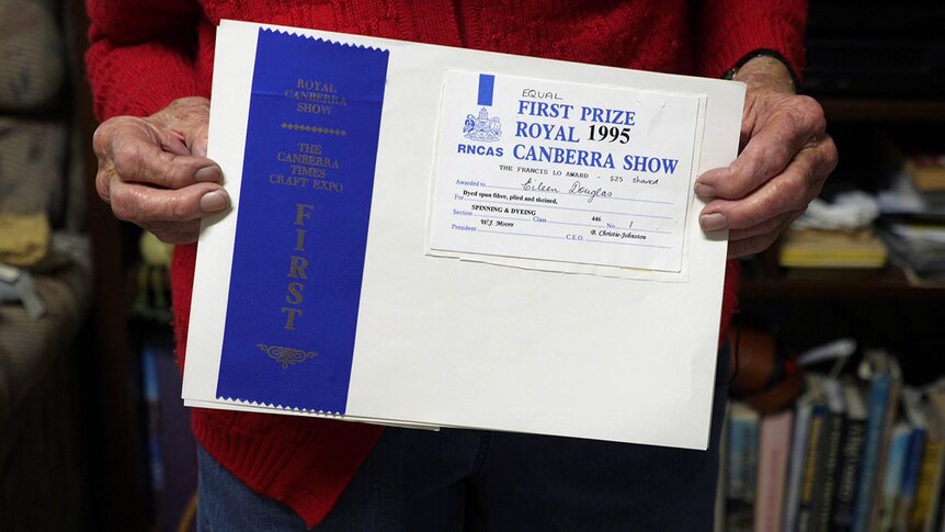 An elderly woman holds a blue first prize ribbon and card mounted on a piece of paper.