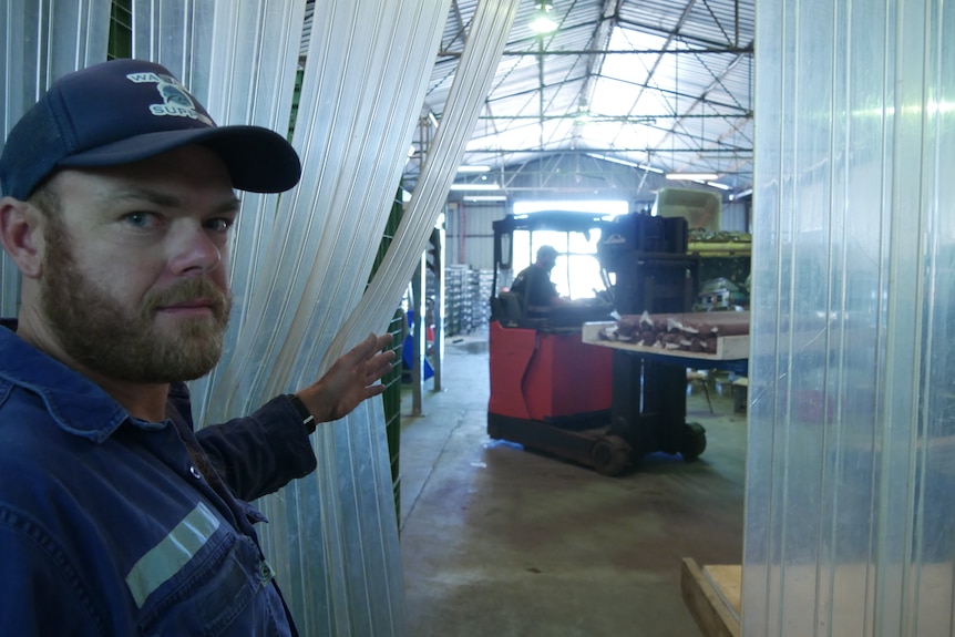 a man holds open plastic blinds, looking onto a factory floor