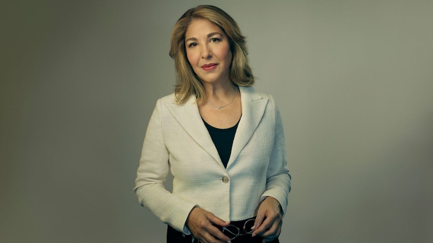 A frontal shot of Naomi Klein wearing a black dress and cream jacket, holding her glasses in her hands