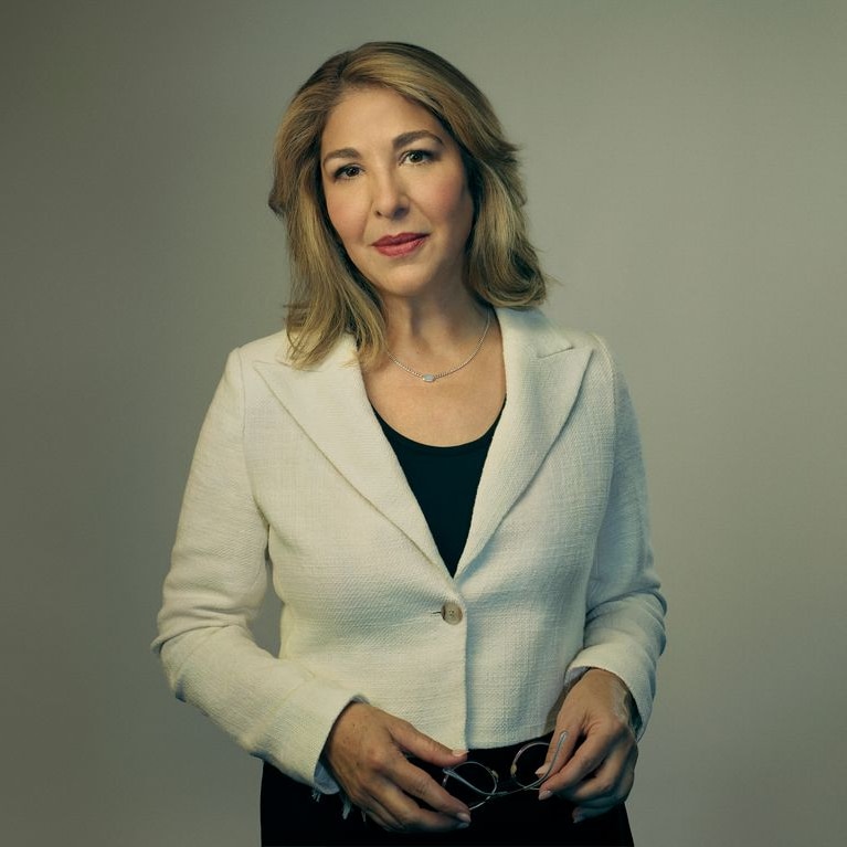A frontal shot of Naomi Klein wearing a black dress and cream jacket, holding her glasses in her hands