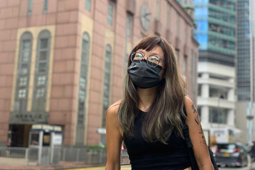 Woman in a black top, wearing a face mask and glasses, standing on a street.