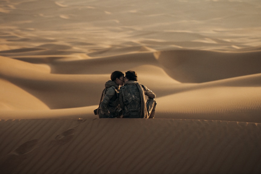Two people in full black suits kiss on top of a sand dune in front of a desert.