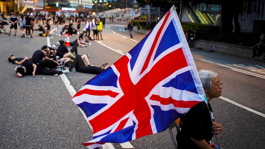 An elderly woman sits down on a chair in the middle of the road while holding Union flag.
